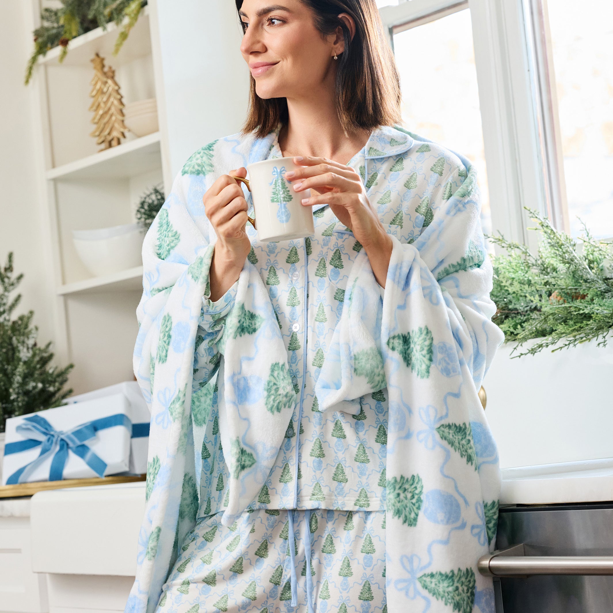 A woman wearing a matching pajama set with a festive Christmas tree and bow print, holding a mug in a bright kitchen setting.