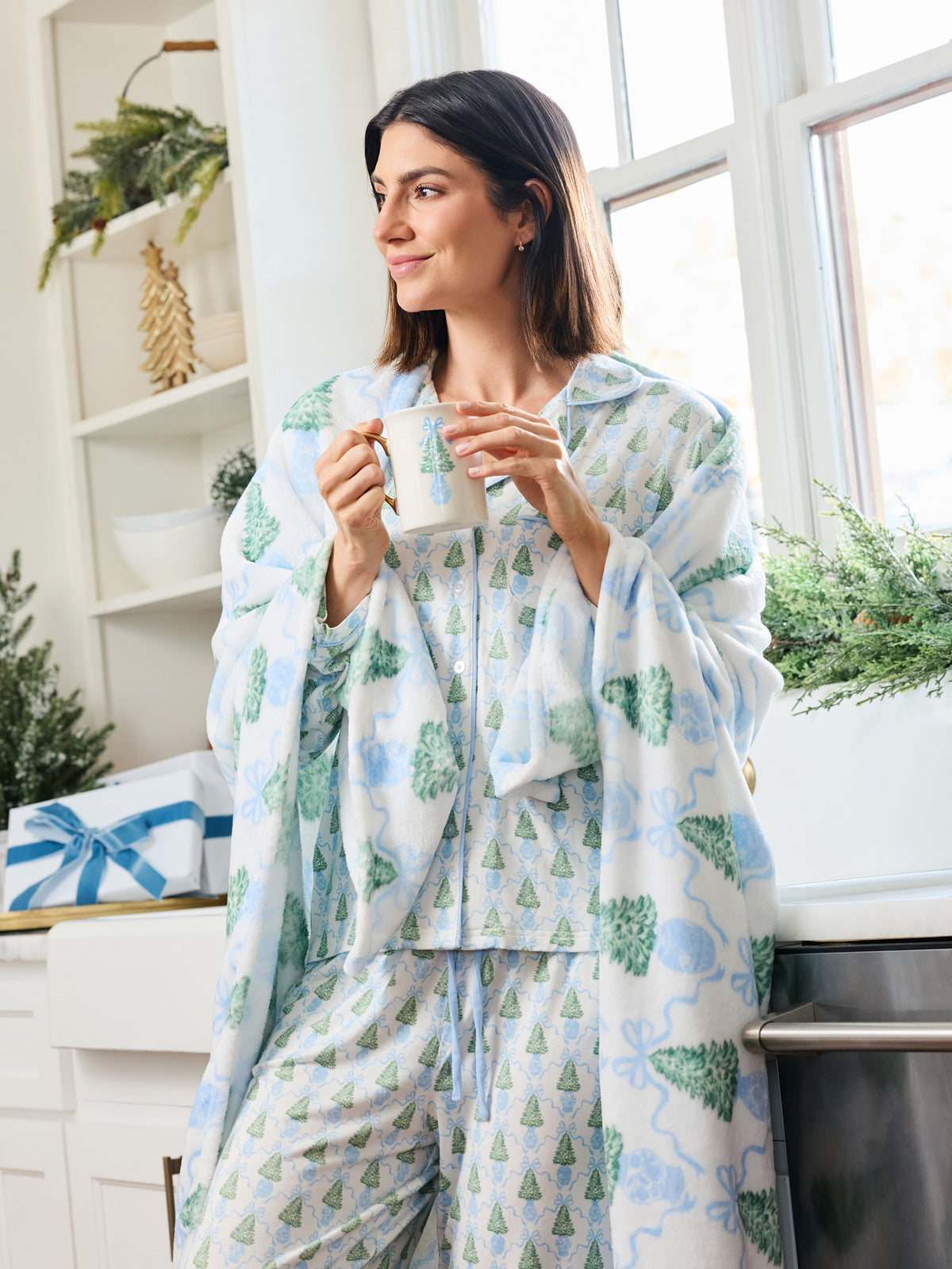A woman wearing a matching pajama set with a festive Christmas tree and bow print, holding a mug in a bright kitchen setting.