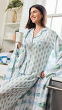 A woman wearing a matching pajama set with a plush throw blanket, featuring a light blue and green Christmas tree pattern, posed in a bright kitchen setting.