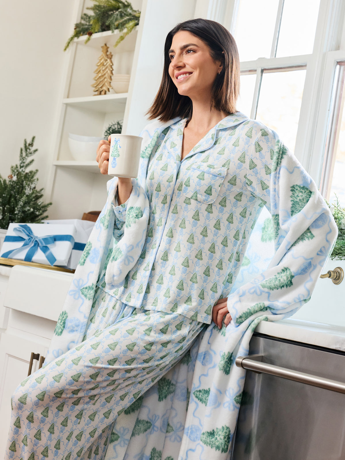 A woman wearing a matching pajama set with a plush throw blanket, featuring a light blue and green Christmas tree pattern, posed in a bright kitchen setting.
