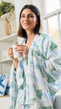 A woman wearing a soft white robe with a light blue and green winter pattern, holding a white mug in a bright, cozy room.