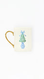 An off-white ceramic mug with a gold handle, featuring a printed illustration of a green Christmas tree in a blue vase with a blue bow on the front.