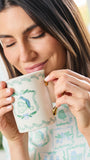 A woman holding a white ceramic mug with a green wreath design and a blue-and-white Santa hat illustration, sipping from it in a bright indoor setting.