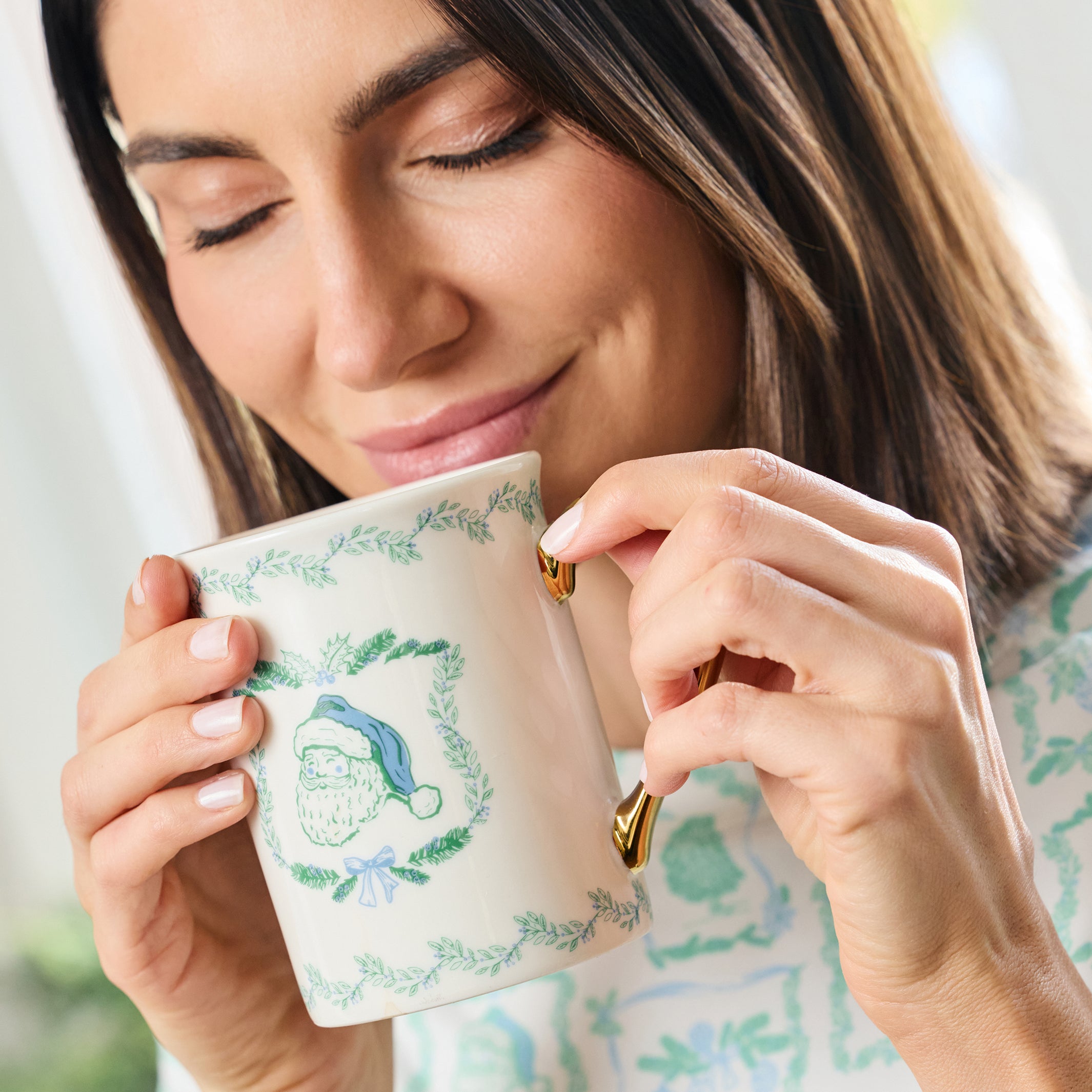 A woman holding a white ceramic mug with a green wreath design and a blue-and-white Santa hat illustration, sipping from it in a bright indoor setting.