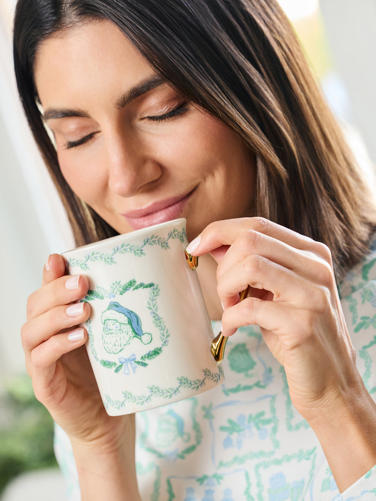 A woman holding a white ceramic mug with a green wreath design and a blue-and-white Santa hat illustration, sipping from it in a bright indoor setting.