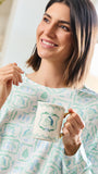 A smiling woman with shoulder-length brown hair holds a white ceramic mug with a blue bird illustration and a gold handle. She is wearing a white long-sleeve top with a light blue botanical pattern in a bright, modern indoor setting.