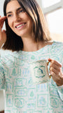 A woman with shoulder-length brown hair smiling while wearing a light green and white patterned long-sleeve top and holding a white ceramic mug with a gold handle.