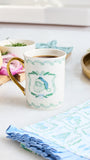 A white ceramic coffee mug with a gold handle and green holiday wreath pattern, filled with coffee on a light tabletop with breakfast items and napkins around it.