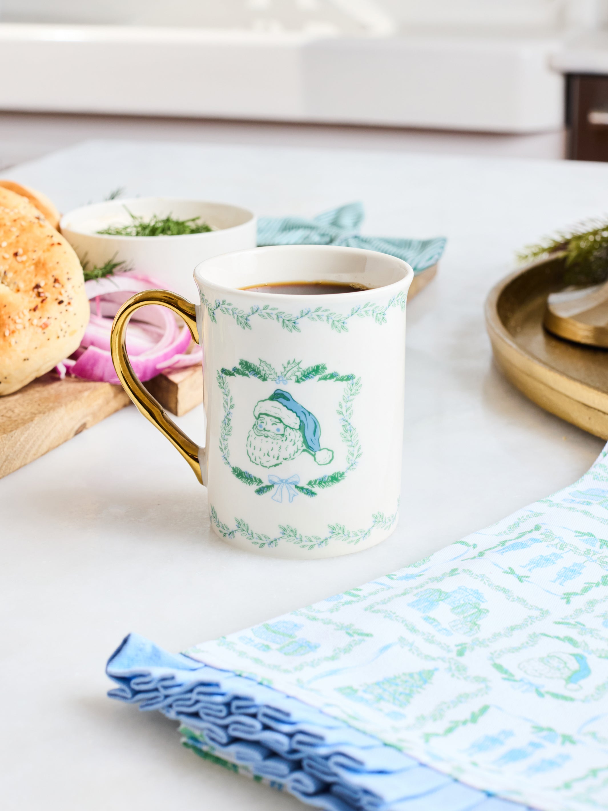 A white ceramic coffee mug with a gold handle and green holiday wreath pattern, filled with coffee on a light tabletop with breakfast items and napkins around it.