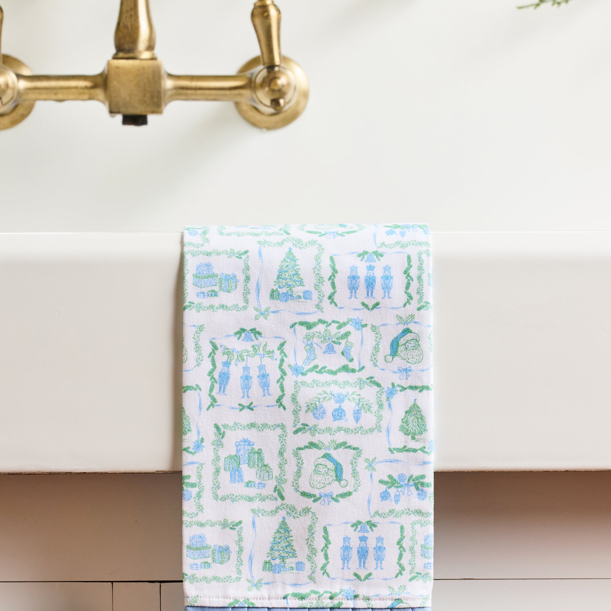 A white kitchen towel with a blue and green winter-themed pattern hangs from a brass faucet spout over a white sink.