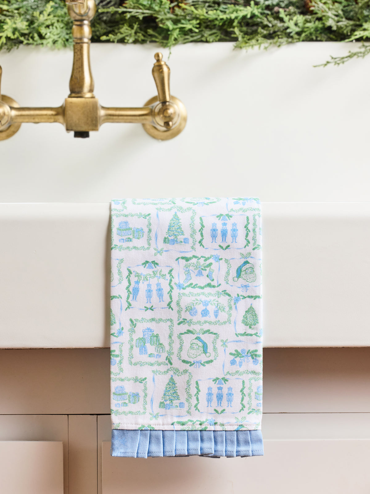 A white kitchen towel with a blue and green winter-themed pattern hangs from a brass faucet spout over a white sink.