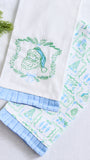 Two white kitchen towels with light blue ruffled edges, one featuring an embroidered green Santa Claus wreath and the other with a green and blue holiday pattern, displayed on a light gray surface with pine branches in the corner.
