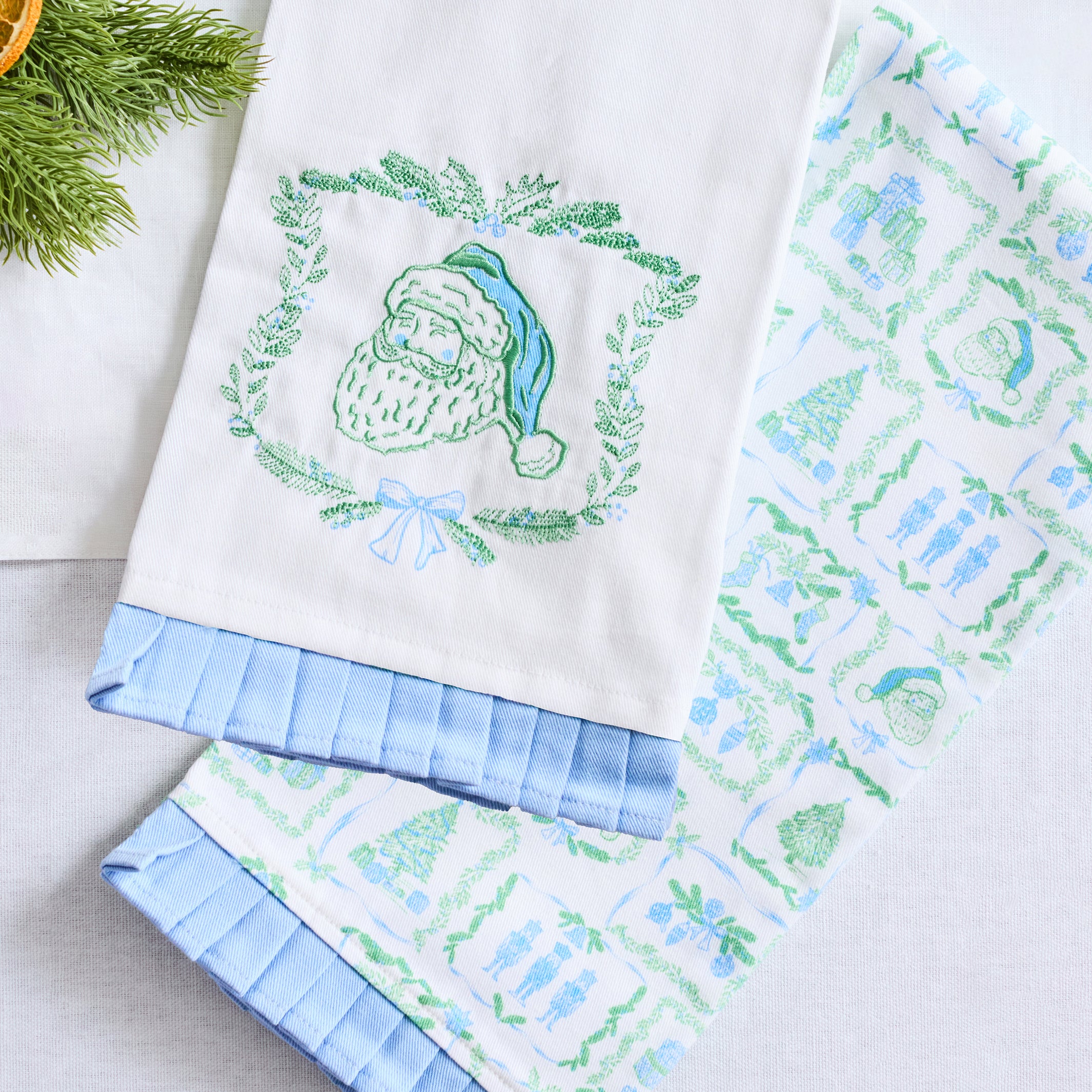 Two white kitchen towels with light blue ruffled edges, one featuring an embroidered green Santa Claus wreath and the other with a green and blue holiday pattern, displayed on a light gray surface with pine branches in the corner.