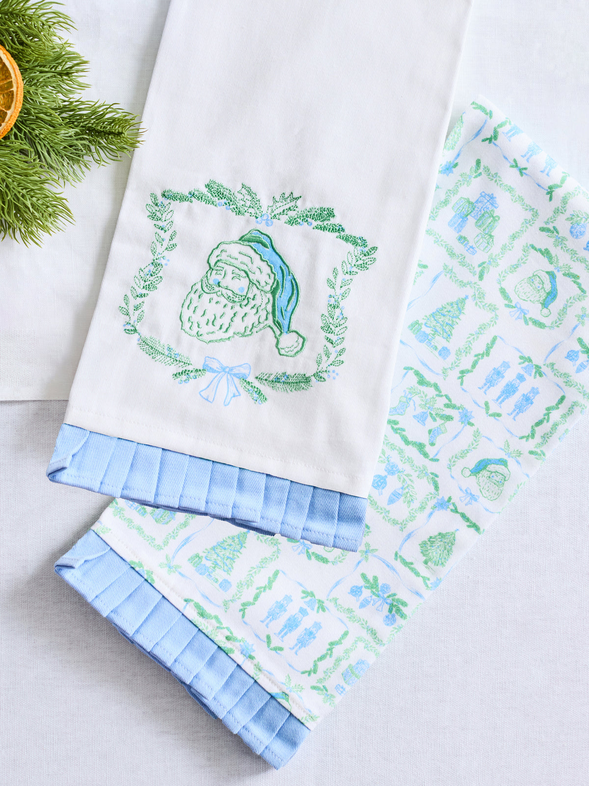 Two white kitchen towels with light blue ruffled edges, one featuring an embroidered green Santa Claus wreath and the other with a green and blue holiday pattern, displayed on a light gray surface with pine branches in the corner.