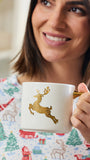 A smiling woman in a Christmas-themed pajama shirt holds a white ceramic mug with a gold glittery reindeer design and a gold handle.