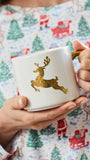 A person holding a white ceramic mug with a gold glittery reindeer design and a gold handle, wearing a festive Christmas pajama shirt with a Santa and tree pattern.