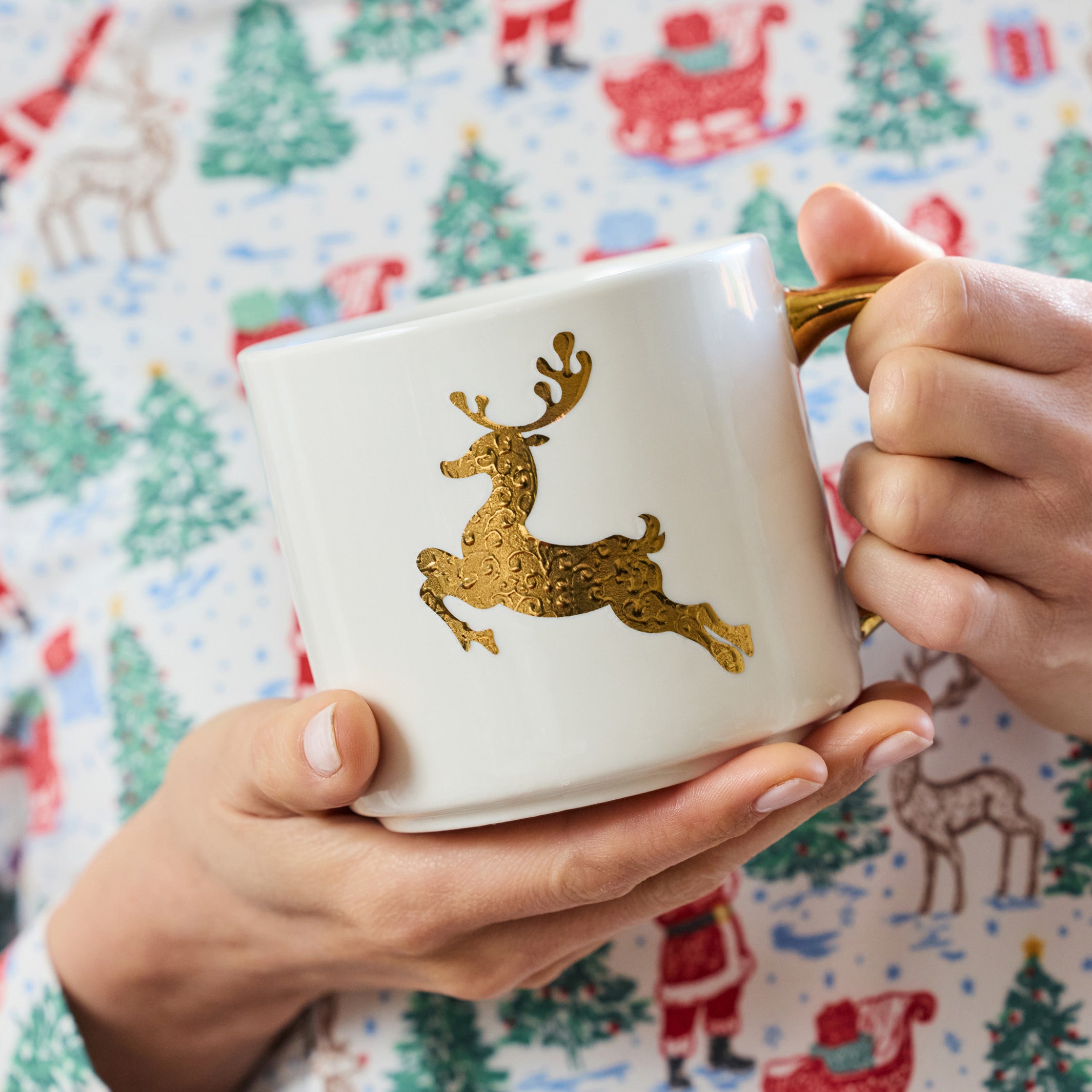 A person holding a white ceramic mug with a gold glittery reindeer design and a gold handle, wearing a festive Christmas pajama shirt with a Santa and tree pattern.