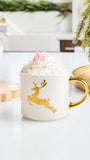 A white ceramic mug with a gold handle and a gold reindeer graphic, filled with whipped cream and topped with pink candy canes, set on a white tabletop with holiday decor in the background.