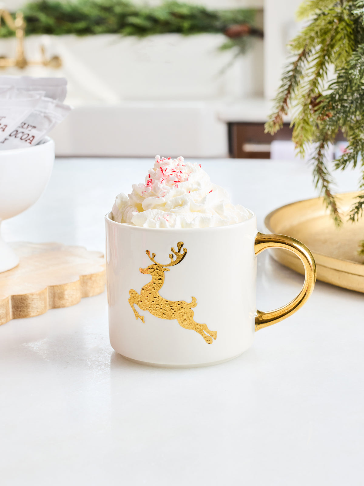A white ceramic mug with a gold handle and a gold reindeer graphic, filled with whipped cream and topped with pink candy canes, set on a white tabletop with holiday decor in the background.