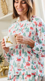 A woman wearing a festive Christmas-themed long-sleeve pajama top and matching pants, holding a white mug with a gold reindeer design. The pajamas feature an all-over print of Santa Claus, reindeer, Christmas trees, sleighs, and gifts on a white background.