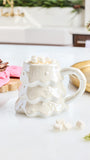 A white ceramic mug shaped like a melting Christmas tree, filled with marshmallows and set on a white countertop in a styled kitchen scene.