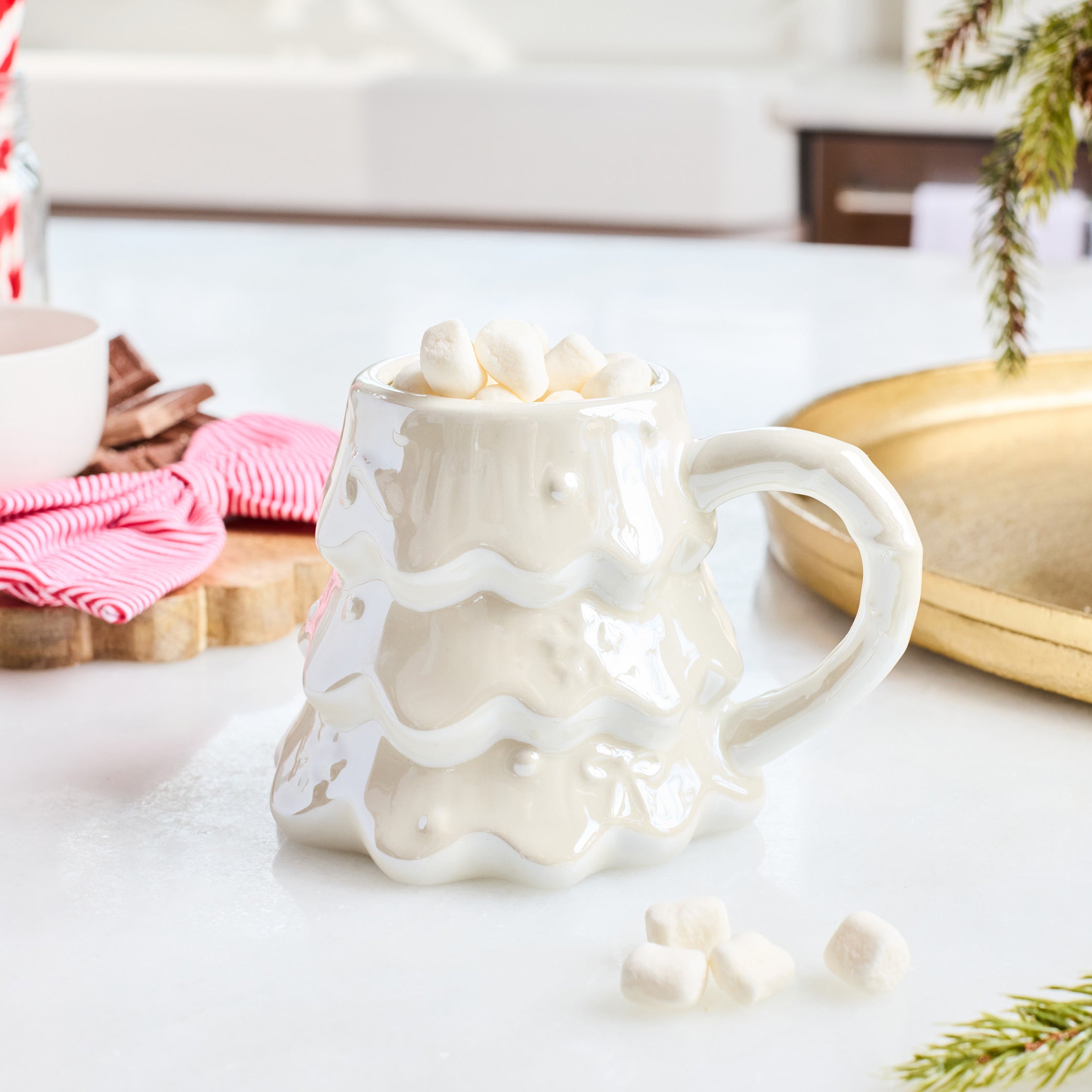 A white ceramic mug shaped like a melting Christmas tree, filled with marshmallows and set on a white countertop in a styled kitchen scene.