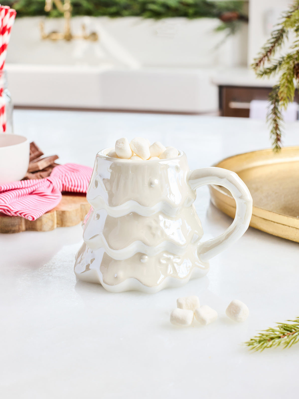 A white ceramic mug shaped like a melting Christmas tree, filled with marshmallows and set on a white countertop in a styled kitchen scene.