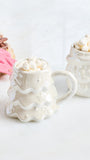 A white ceramic mug shaped like a Christmas tree, filled with mini marshmallows, alongside a matching white ceramic container on a light surface with holiday-themed props in the background.