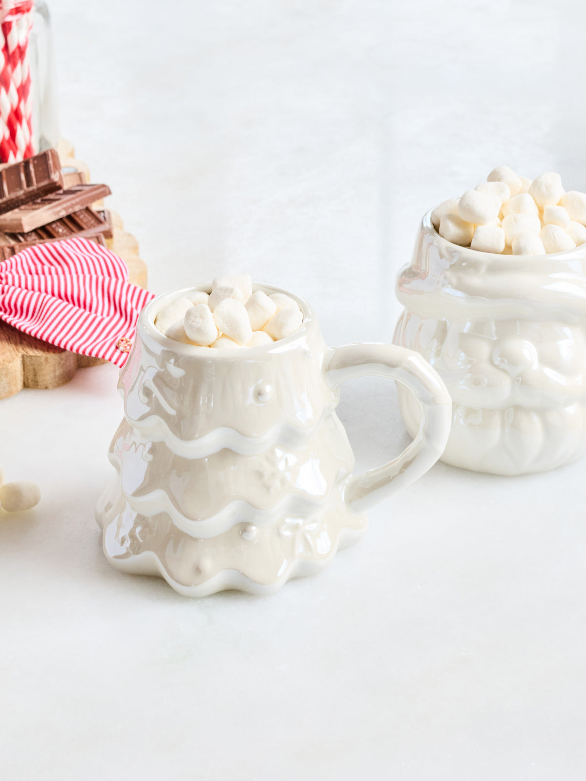 A white ceramic mug shaped like a Christmas tree, filled with mini marshmallows, alongside a matching white ceramic container on a light surface with holiday-themed props in the background.