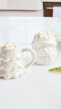 Two white ceramic mugs shaped like a Christmas tree and Santa's sack, both filled with marshmallows, set on a white marble countertop with holiday decorations.
