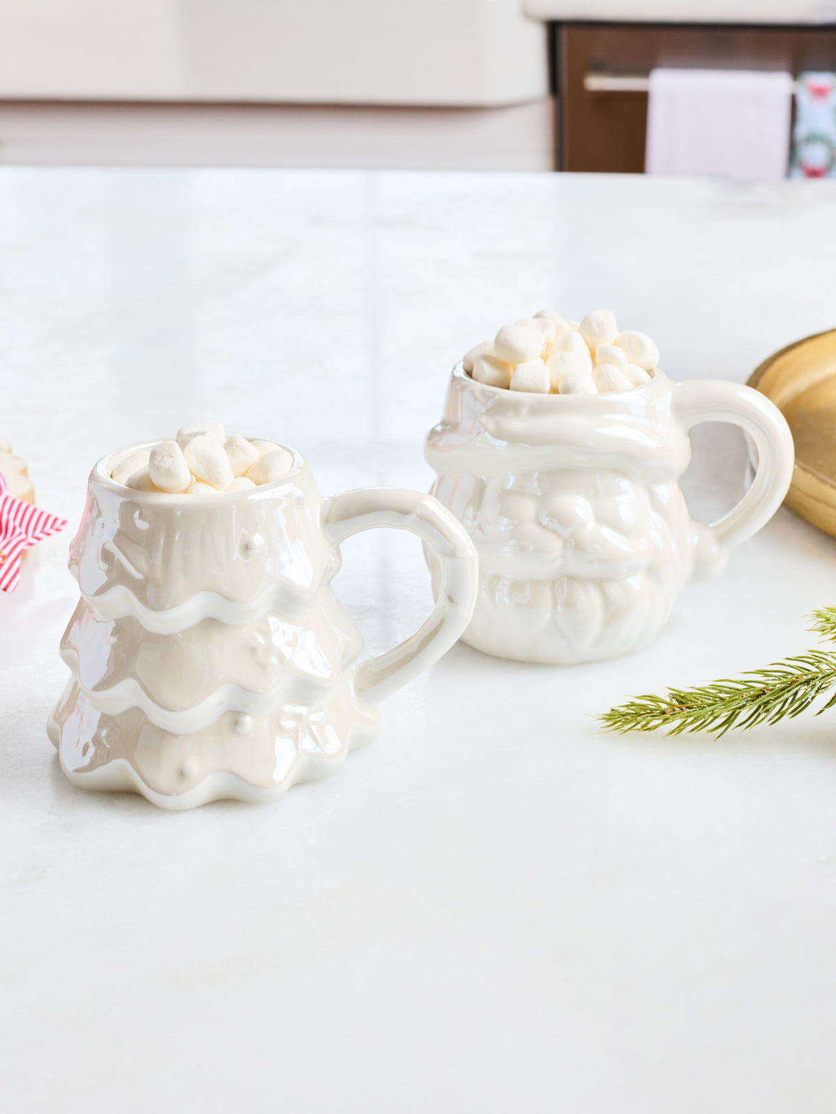 Two white ceramic mugs shaped like a Christmas tree and Santa's sack, both filled with marshmallows, set on a white marble countertop with holiday decorations.