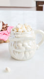 A white ceramic Santa mug filled with mini marshmallows, set on a white countertop with a pink striped napkin and a small bowl in the background.
