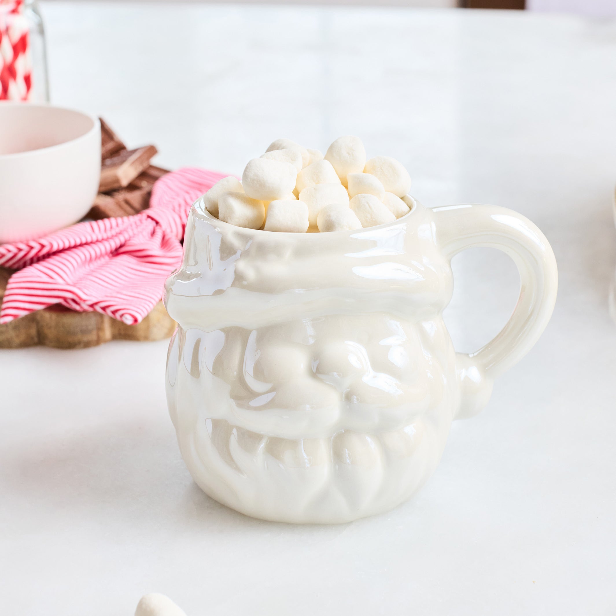 A white ceramic Santa mug filled with mini marshmallows, set on a white countertop with a pink striped napkin and a small bowl in the background.