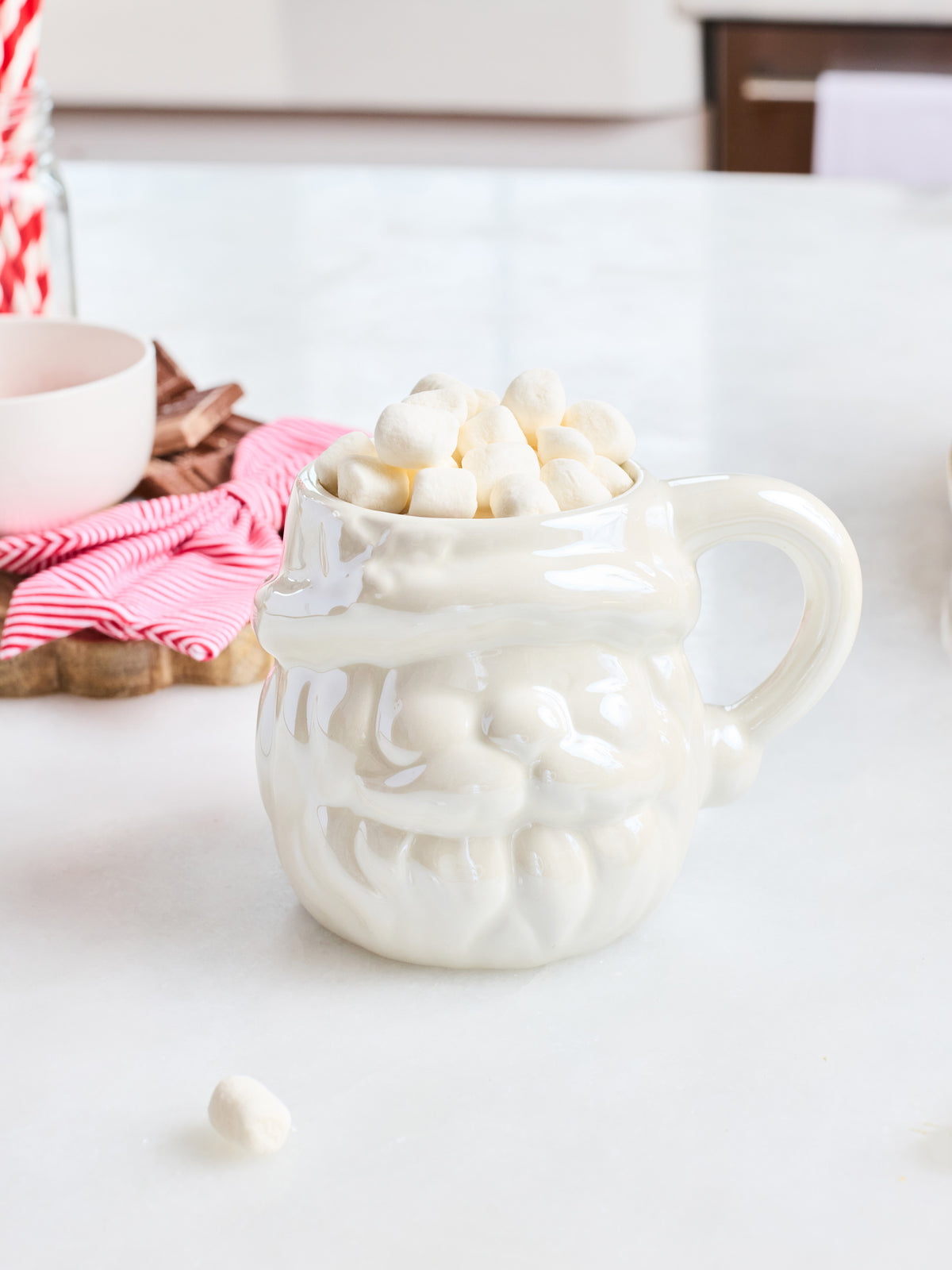 A white ceramic Santa mug filled with mini marshmallows, set on a white countertop with a pink striped napkin and a small bowl in the background.