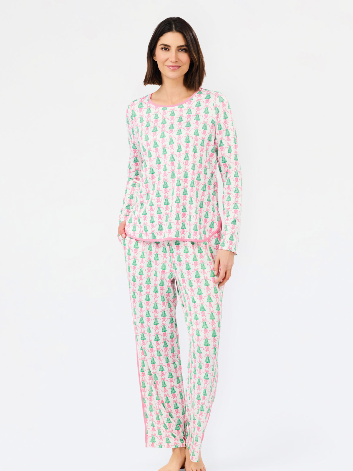 A woman modeling a two-piece long-sleeve pajama set with a white background and an all-over green pine tree pattern, accented with pink piping on the collar, cuffs, and waist, and pink side stripes on the pants.