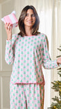 A woman wearing a pink and mint green pajama set with a repeating Christmas tree pattern, holding a pink gift box next to a lit Christmas tree.