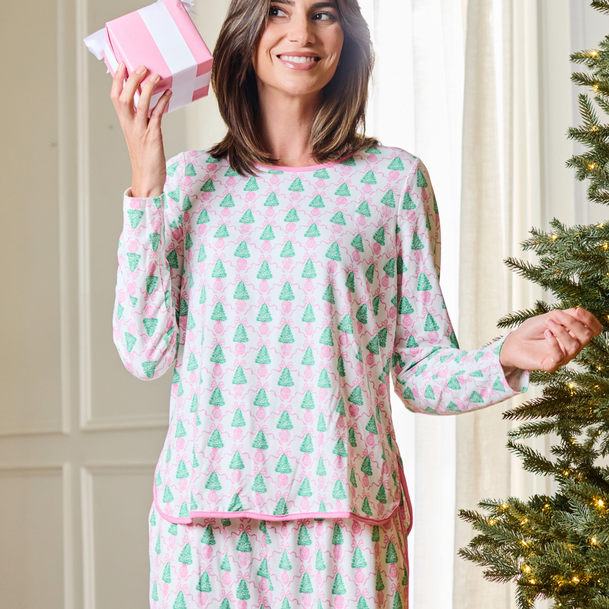A woman wearing a pink and mint green pajama set with a repeating Christmas tree pattern, holding a pink gift box next to a lit Christmas tree.