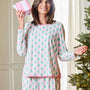 A woman wearing a pink and mint green pajama set with a repeating Christmas tree pattern, holding a pink gift box next to a lit Christmas tree.
