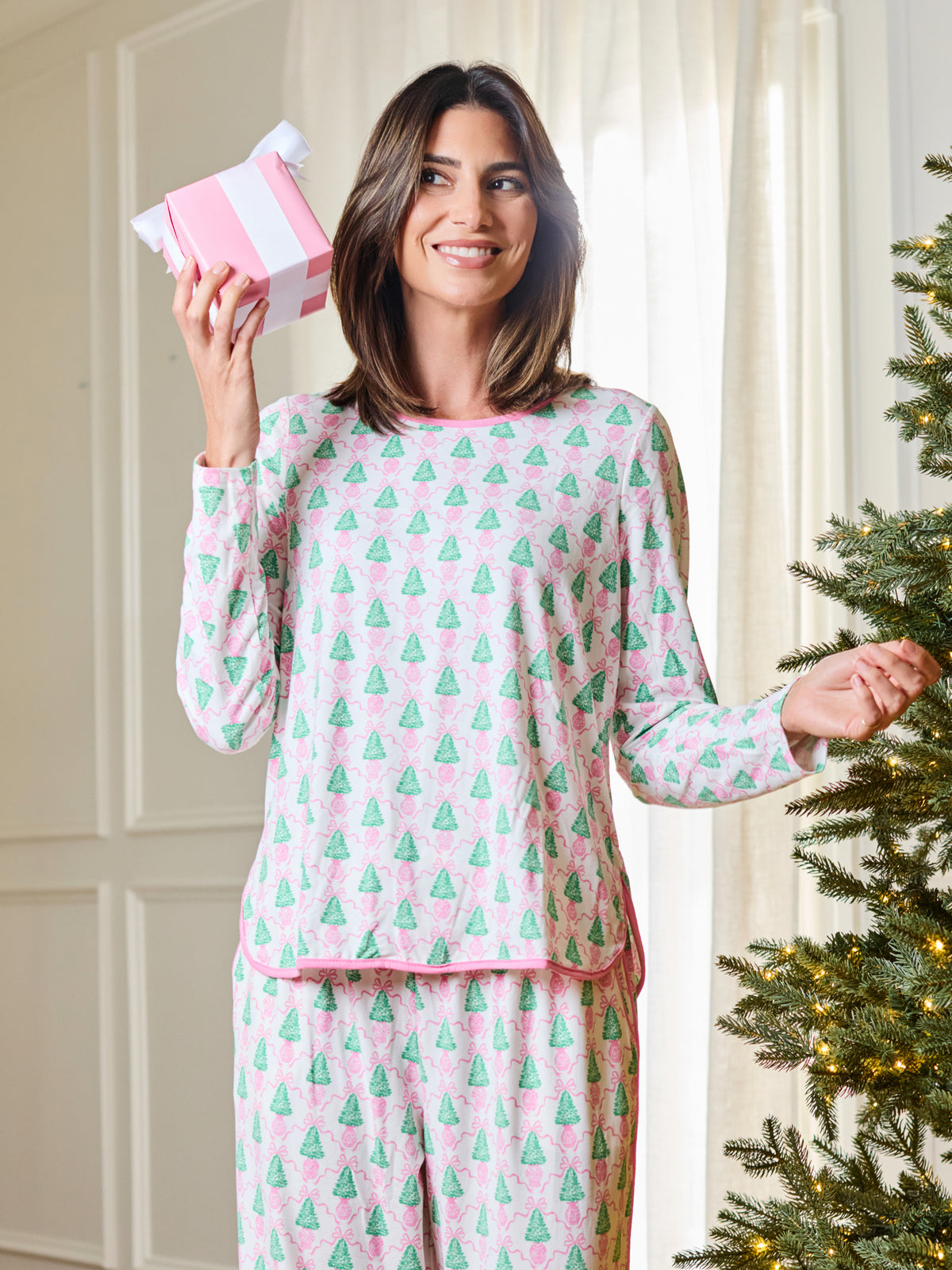 A woman wearing a pink and mint green pajama set with a repeating Christmas tree pattern, holding a pink gift box next to a lit Christmas tree.
