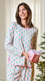 A woman wearing a pink and green Christmas tree patterned pajama set holds a pink gift box with a white bow, standing next to a decorated Christmas tree in a bright indoor setting.