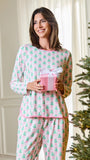 A woman wearing a pink and green Christmas-themed pajama set stands indoors next to a decorated Christmas tree, holding a pink gift box with a white bow.
