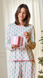 A woman wearing a pink and green Christmas tree patterned pajama set holds a pink gift box with a white bow, standing in a warmly lit room next to a decorated Christmas tree.