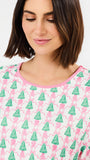 A woman wearing a white long-sleeve top with a festive Christmas print of green pine trees, pink ornaments, and pink wavy lines, featuring a pink crew neckline.
