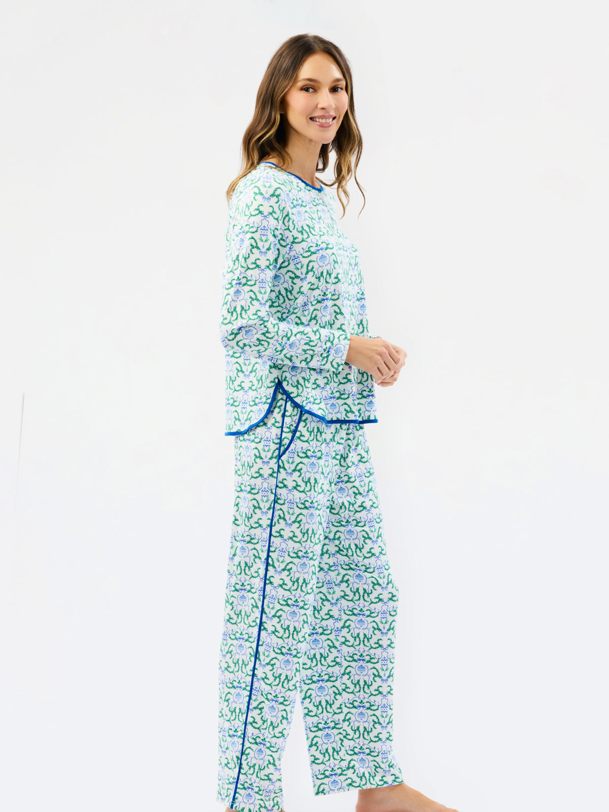 A woman modeling a matching two-piece holiday pajama set with a green and blue floral pattern and blue trim, standing against a plain white studio background.