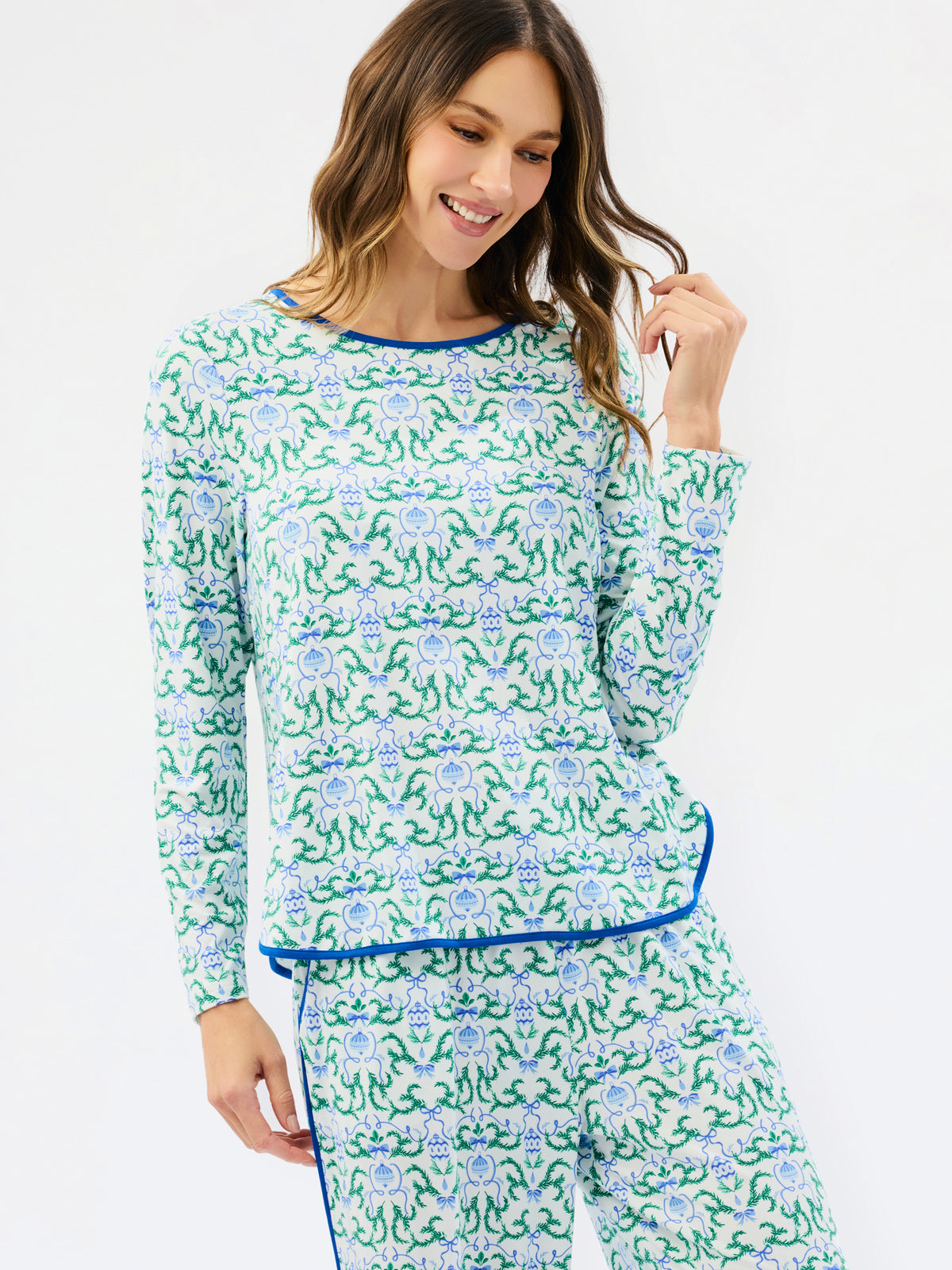 A woman modeling a matching long-sleeve pajama top and pants set with a green and blue floral pattern and blue trim, against a plain white studio background.