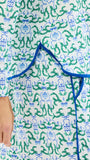A close-up of a white fabric garment with an all-over green and blue botanical print, featuring a blue tie closure at the waist and a hand visible on the left side.