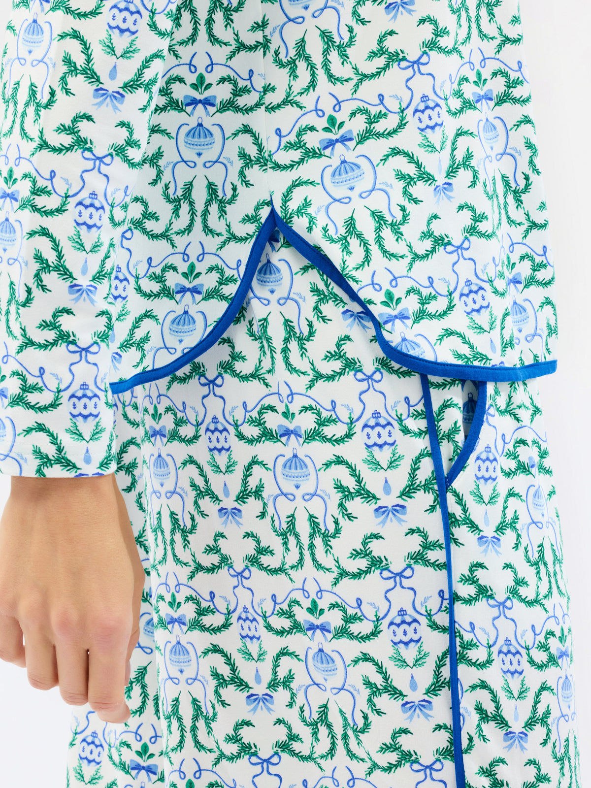A close-up of a white fabric garment with an all-over green and blue botanical print, featuring a blue tie closure at the waist and a hand visible on the left side.