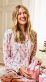 A smiling blonde woman sits cross-legged indoors wearing a light pink pajama set with a colorful Christmas-themed print. She is holding a pink gift box with a white bow, with a Christmas tree visible at the right edge.
