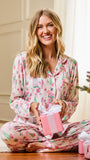 A woman wearing pink holiday-themed pajamas sits cross-legged on a wooden floor, smiling while holding a pink gift box with white ribbon. A Christmas tree is visible on the right, and other wrapped presents are on the floor nearby.