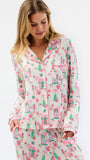 A woman wearing a matching long-sleeve pajama top and pants set with a whimsical pink and green house pattern on a cream background, photographed against a plain white studio backdrop.